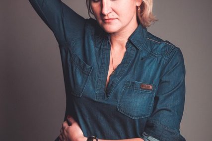 Woman with short blonde hair poses in a denim shirt against a neutral background.