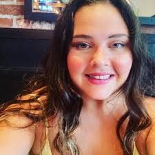 Shea Gaier, CEO Smiling woman with long, wavy brown hair wearing a spaghetti strap top. There is a picture on the red and white brick wall behind her.