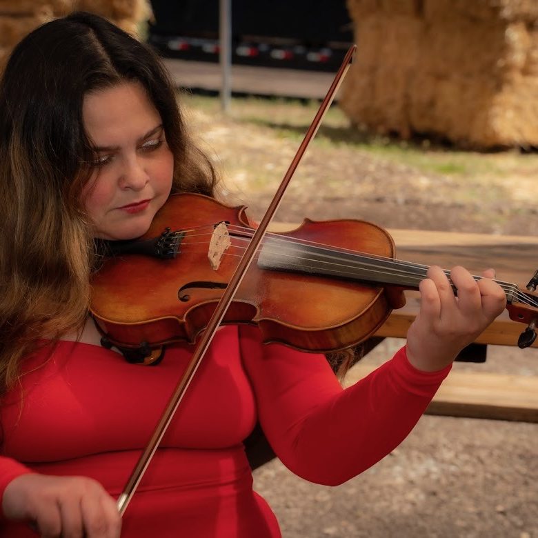 White woman wearing a long sleeved top. She has long, wavy brown hair with highlights and she is holding a violin/fiddle