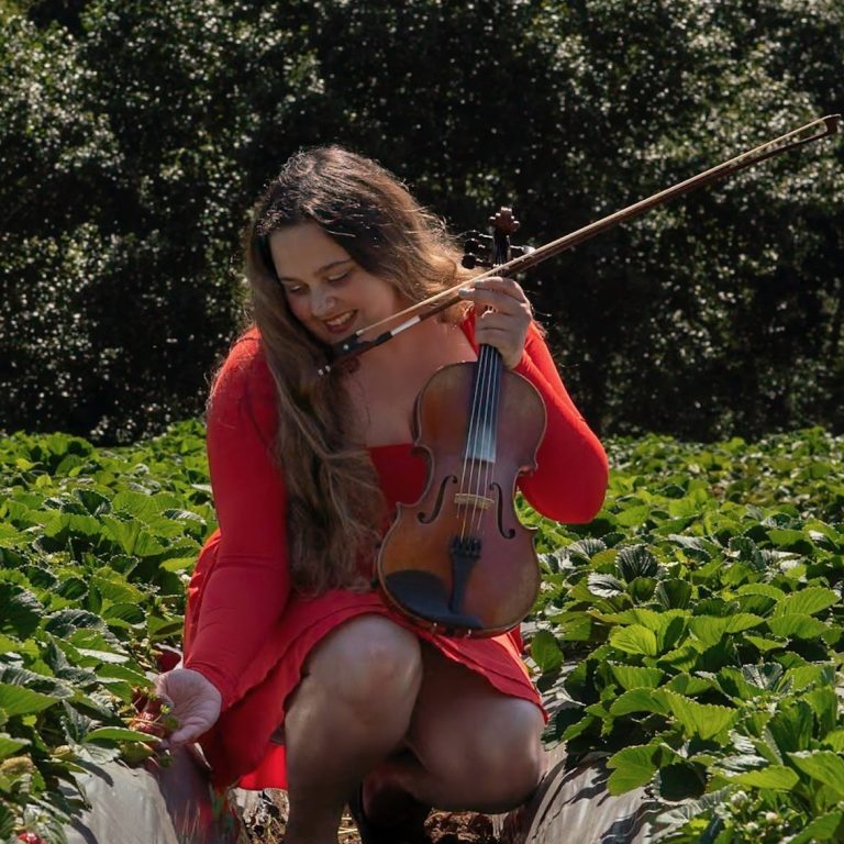 A woman in a red long sleeved dress holding a violin among green plants.