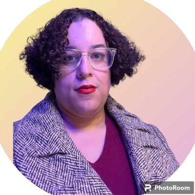 Woman with short, curly hair and glasses wearing a maroon v-neck shirt and a patterned coat. She is also wearing a red lipstick.