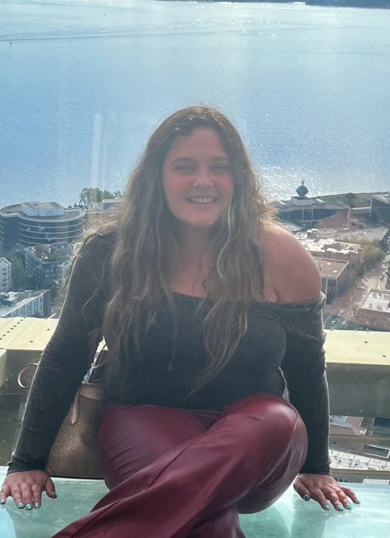 A woman with long hair seated overlooking a cityscape. She is wearing maroon pants and a black shirt which has one long sleeve and one short